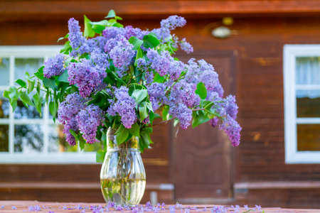A bouquet of beautiful lilacs in a glass vase stands on a wooden table. Wooden cottage in the forest.の写真素材