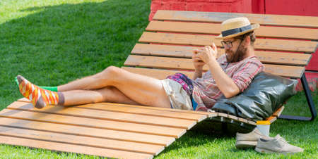 Moscow. Russia. August 14 2021. A hipster with glasses and a hat looks at the phone on a sunny summer day.のeditorial素材