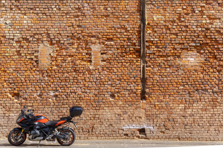 Black and orange motorcycle parked on asphalt in front of a brick wall with cracks and weathered details. The contrast between the vehicle and aged facade creates a striking composition.の写真素材