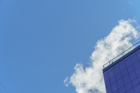 Corner of a modern building with reflective glass windows against a clear blue sky with a light cloud. Diagonal composition emphasizes the contrast between architecture and nature.の写真素材