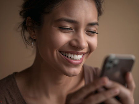 A woman with tied-back hair smiles while looking at her smartphone. The neutral background highlights her expression and the moment of digital connection.の素材