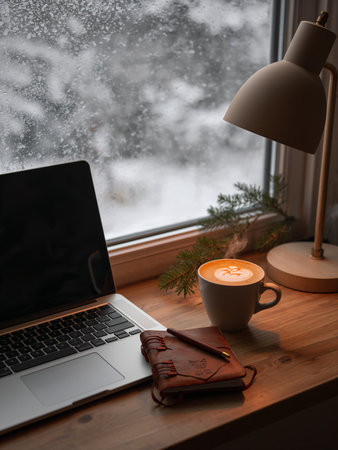 A cozy winter workspace with a laptop, a warm coffee, and festive decor overlooks a snowy window view. This inviting scene creates a perfect atmosphere for working from home during the holidays.の素材