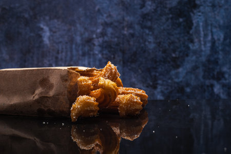 Sugared churros on black table with reflection. Churros in paper bag. Blue background.