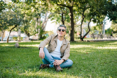 Portrait of gray-haired man wearing sunglasses in a park sitting on the grass looking at the camera.の写真素材