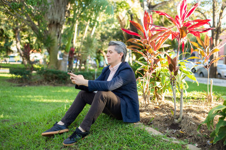Gray-haired man looking at his cell phone sitting on the grass in a park.の写真素材