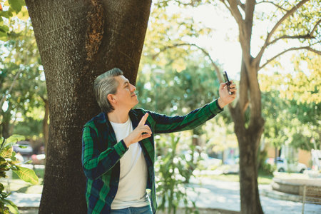 Portrait of gray haired man in a park taking a selfie close to a tree.の写真素材