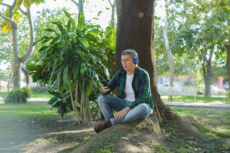 Gray-haired man listening to music with headphones and typing on his cell phone sitting in a park next to a tree.の写真素材