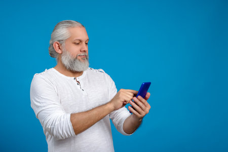 Man with gray hair and beard typing on his phone. blue backgroundの写真素材
