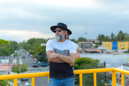 Long-haired, gray-haired man wearing a black hat on a pedestrian bridge overlooking the city.の写真素材