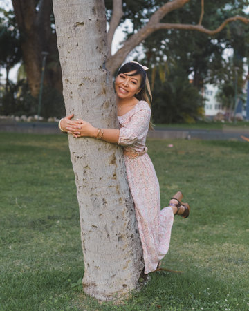 Woman wearing dress and hair bow hugging a tree in a public park. Positive emotion.の写真素材