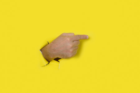 Male hand coming out of a hole in a yellow torn paper background pointing to the right.の写真素材