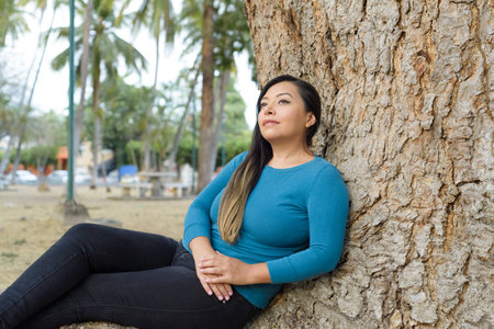 Woman seated leaning against a tree with pensive attitude.
