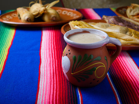 Clay cup with atole over colorful serape. Tamales in the background. Mexican food.の写真素材