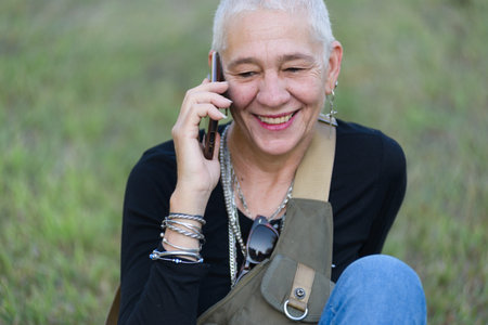 Middle-aged woman smiles sitting on the grass in the park while talking on a cell phone.