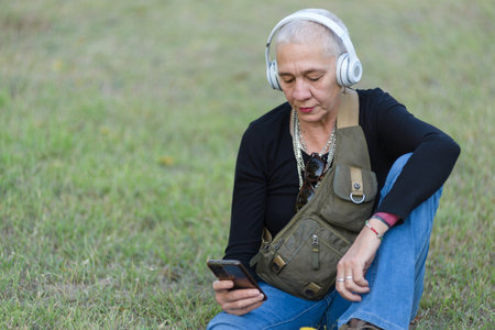 Middle-aged woman sitting on the grass in the park while listening to music with headphones and looking at her cell phone.の写真素材