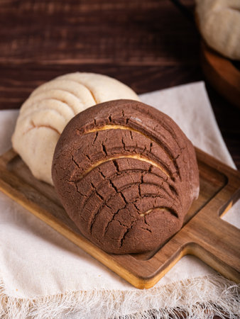 Close-up of two concha breads, one chocolate and one vanilla, resting on a wooden board.