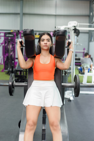A young woman intensely focuses on a shoulder press exercise using a weight machine at the gym.の写真素材