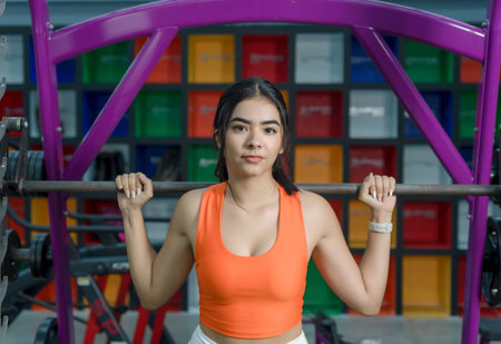 A young woman, wearing an orange sports bra, confidently holds a barbell in a gym setting.の写真素材