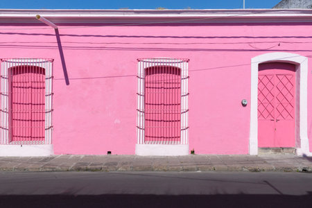 A sunlit pink building with three pink doors, two with metal grills, stands on a quiet street.