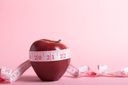 A red apple encircled by a measuring tape rests on a pink background.