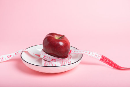 A vibrant red apple on a white plate encircled by a pink measuring tape. Diet, health, and weight...