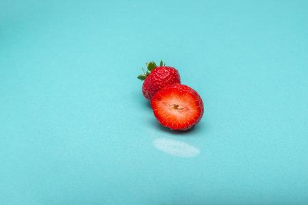 A whole and a halved strawberry resting on a smooth turquoise background.