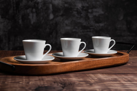 Three white espresso cups rest on matching saucers, arranged neatly on a rustic wooden serving trayの写真素材