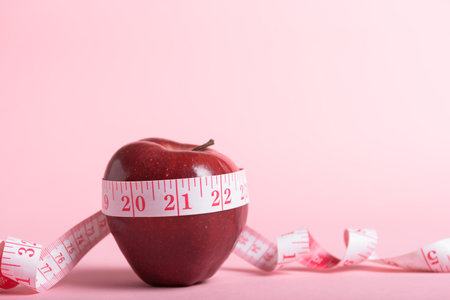 Red apple and a pink measuring tape on a pink background, healthy eating and weight management.