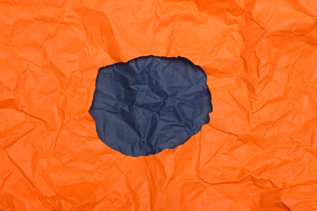 A circular opening in crumpled orange paper, revealing a background of dark blue, similarly creased...