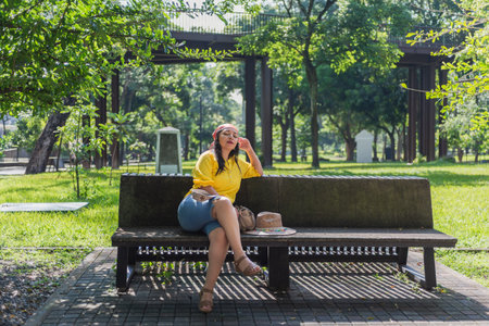 A woman in a yellow shirt and denim shorts sits on a park bench, enjoying the shade. A hat and bag...