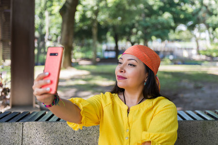 A young woman in a yellow shirt and orange headscarf sits on a park bench, taking a selfie with her...