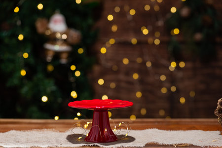 A vibrant red cake stand sits on a rustic wooden table, adorned with warm fairy lights against a blurred Christmas backdrop.の写真素材