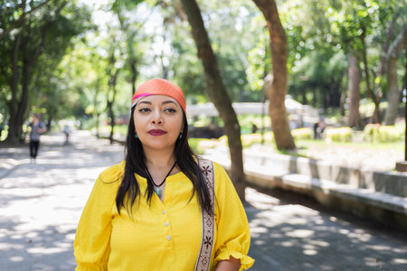 A woman with long dark hair and an orange headscarf stands confidently in a park, wearing a bright yellow top.の写真素材