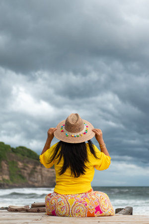 Back view of a woman wearing a straw hat is sitting on a wooden pier, looking out at the ocean under a dramatic, cloudy sky.の写真素材