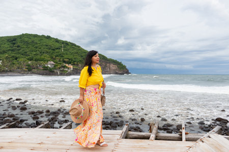 A woman stands on a wooden pier, looking out towards the ocean on a cloudy day.