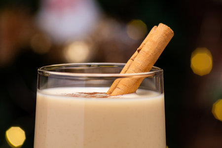 Traditional Holiday Drink Eggnog with Cinnamon Stick set against a blurred background of Christmas lights and decorationsの写真素材