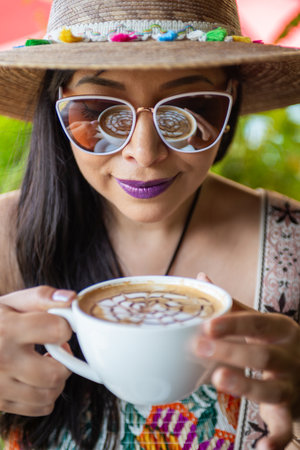 A close up image of a smiling woman wearing a hat and sunglasses holding a cup of latte art drink.の写真素材