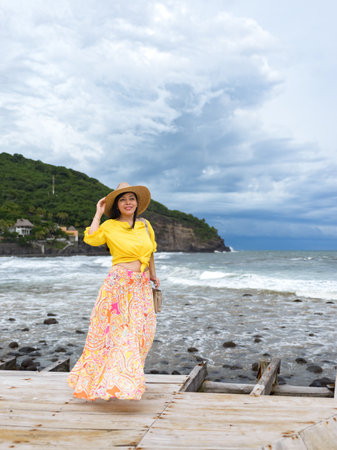 A woman with a straw hat and long skirt standing on a wooden pier with a view of the sea.の写真素材
