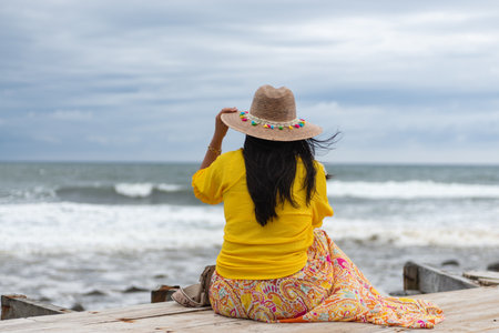 A woman sits on a wooden boardwalk, gazing out at the ocean, wearing a hat, yellow top, and patterned skirtの写真素材