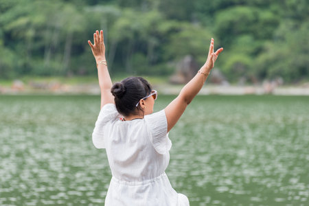 A serene image shows the back of a woman in a white dress with her arms raised, meditating by a calm...
