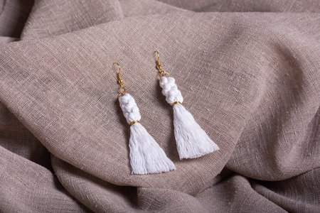 A pair of white macrame tassel earrings with golden accents resting on textured beige fabric.の写真素材
