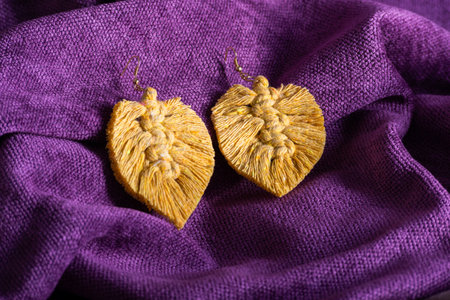 Close-up of handmade macrame leaf earrings in a yellow-gold color, displayed on a purple fabric background.の写真素材