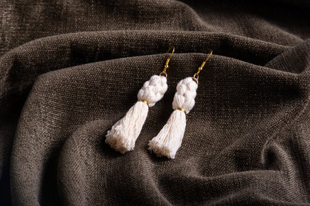 A pair of handmade macrame tassel earrings featuring intricate knotting and golden ring accents, displayed on a richly textured, draped fabric background.の写真素材