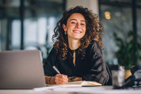 A cheerful, curly-haired woman smiles as she works at her desk with a laptop, notebook, and pen in an office environment.の素材