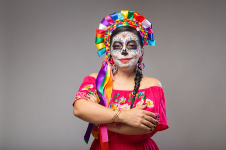 Portrait of a woman with colorful Catrina makeup standing with crossed arms against a gray background.