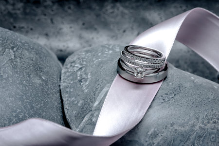 The bride and groom's wedding rings sit on top of a pink satin ribbon laying across grey stone carved hearts.の写真素材