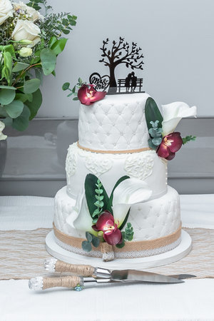 A beautiful three tiered wedding cake adordned with rose petals, greenery and rustic burlap ribbon with the cutting knife and server utensils.の写真素材