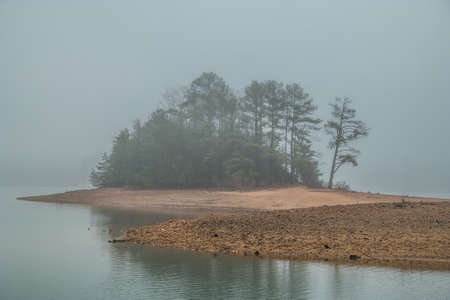 Thick morning fog at the lake with the woodlands on a small island obscured by the conditions of the weather in wintertimeの写真素材