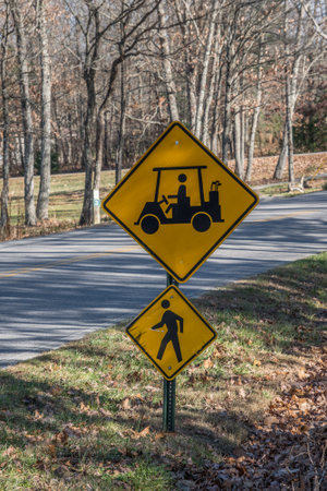 Two signs on a pole alongside the street by a golf course and trails showing a golf cart and pedestrian crossing ahead to approach with caution closeup viewの写真素材