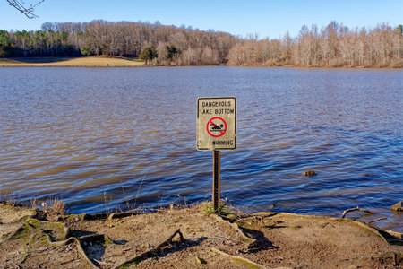 Old grungy signage along the shoreline at the lake saying dangerous lake bottom no swimming which is full of rocks and boulders closeup view on a sunny day in late wintertimeの写真素材